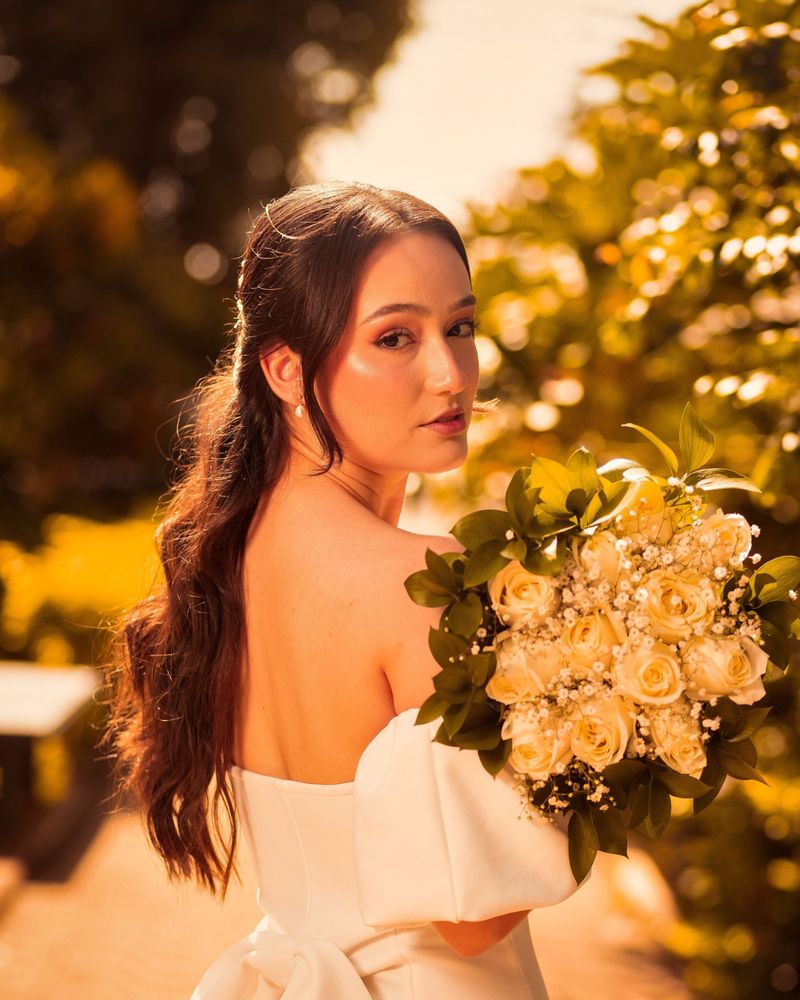Bride in an off-the-shoulder gown glancing back over her shoulder in golden sunlight, with a soft glowing makeup look and a bouquet of cream roses