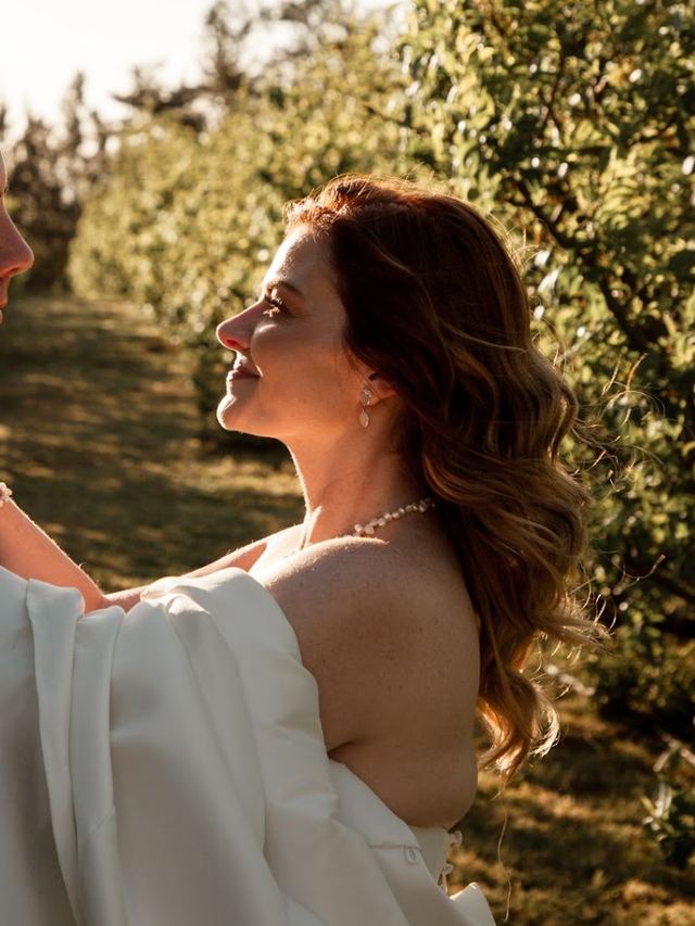 Bride with long caramel waves gazing at her groom in an orchard at sunset, pearl jewellery
