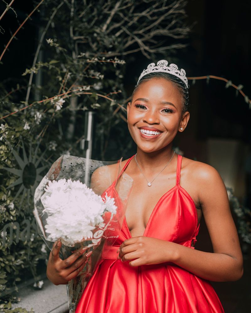 Smiling girl in a red satin dress with a silver tiara, holding a bouquet of white flowers