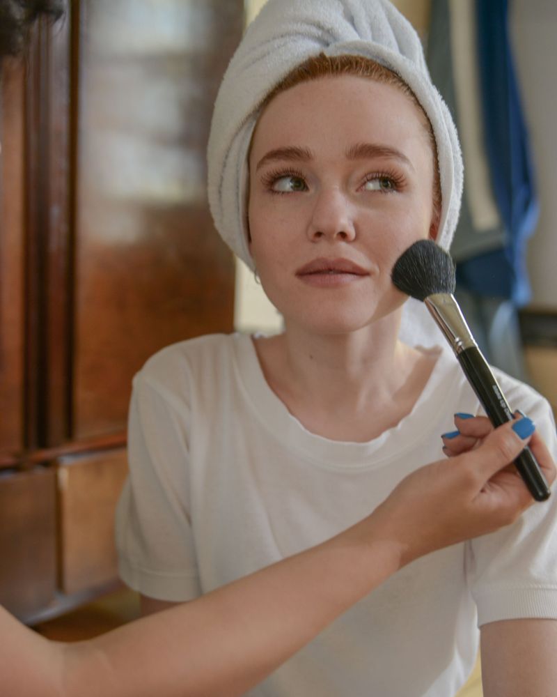 Woman with a white towel wrapped around her hair looking up while a makeup artist brushes blush onto her cheek