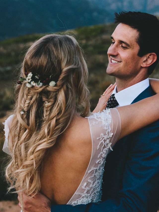 Bride with a loose fishtail half-up hairstyle and wildflowers in her hair, embracing her groom in the mountains