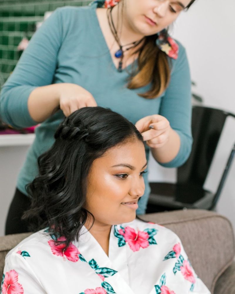 Makeup artist pinning a bride's half-up curled hair while the bride sits in a floral robe with a soft glowing makeup look