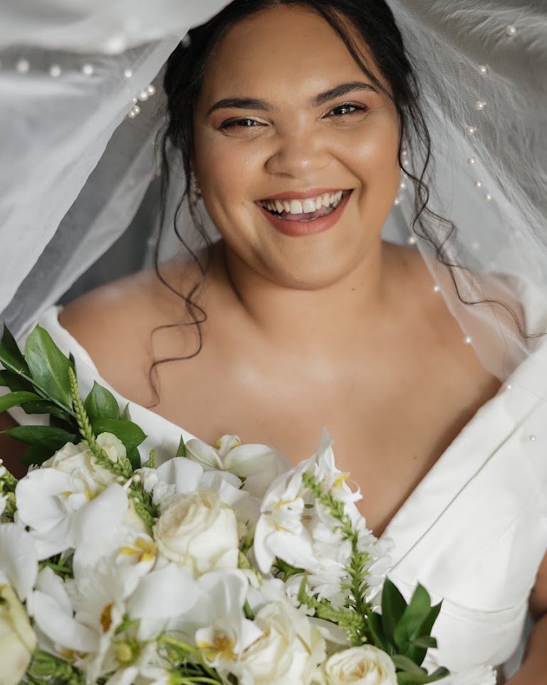 Smiling bride under a pearl-trimmed veil, holding a bouquet of white roses and orchids