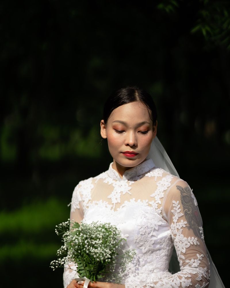 Bride with sleek centre-parted hair in a lace gown looking down, soft peach eyeshadow and a red lip, holding a baby's breath bouquet