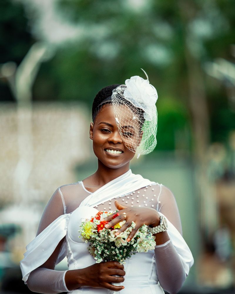 Smiling bride wearing a birdcage veil with a white flower, natural glowing makeup, holding a small wildflower bouquet