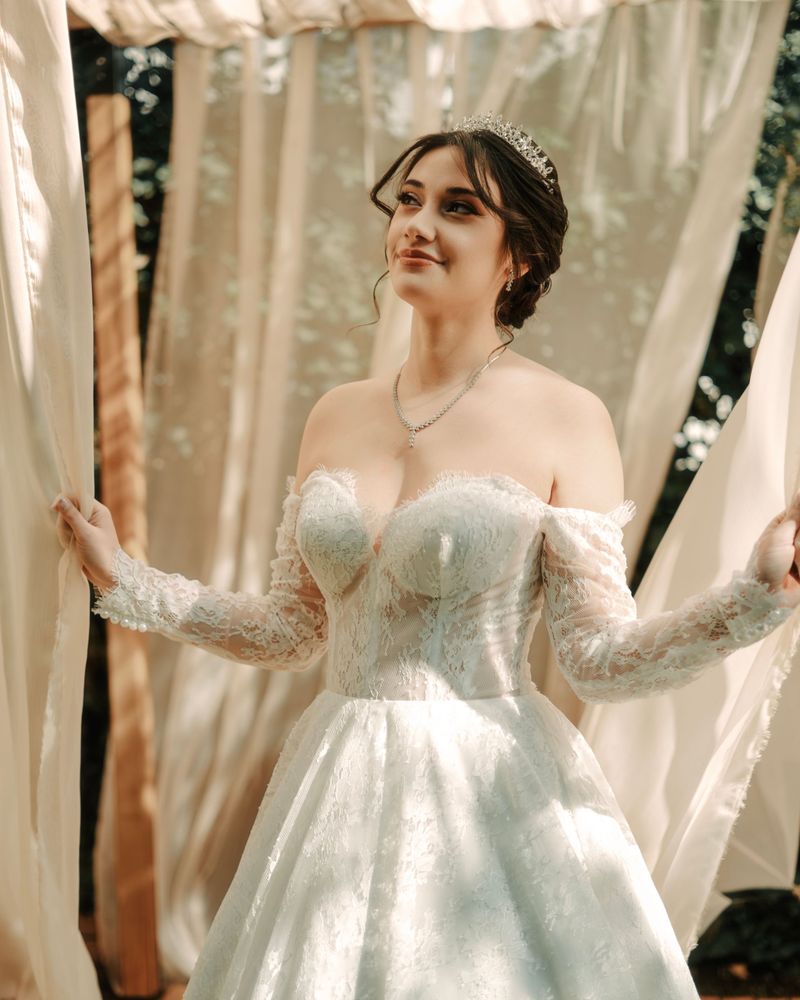 Smiling bride in a lace wedding gown and silver tiara standing between soft cream curtains