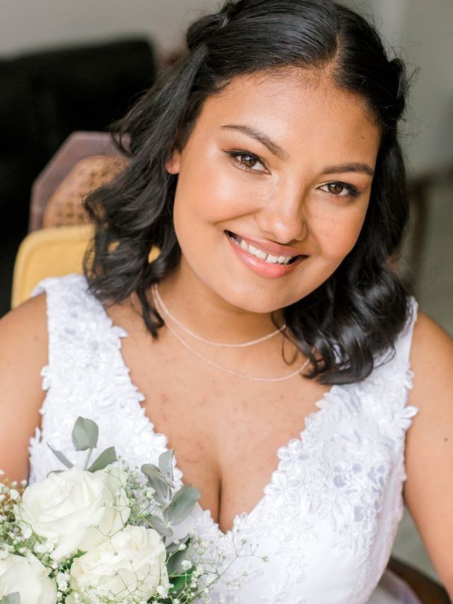 Bride with shoulder-length curls and a glowing peach makeup look, holding white roses and baby's breath