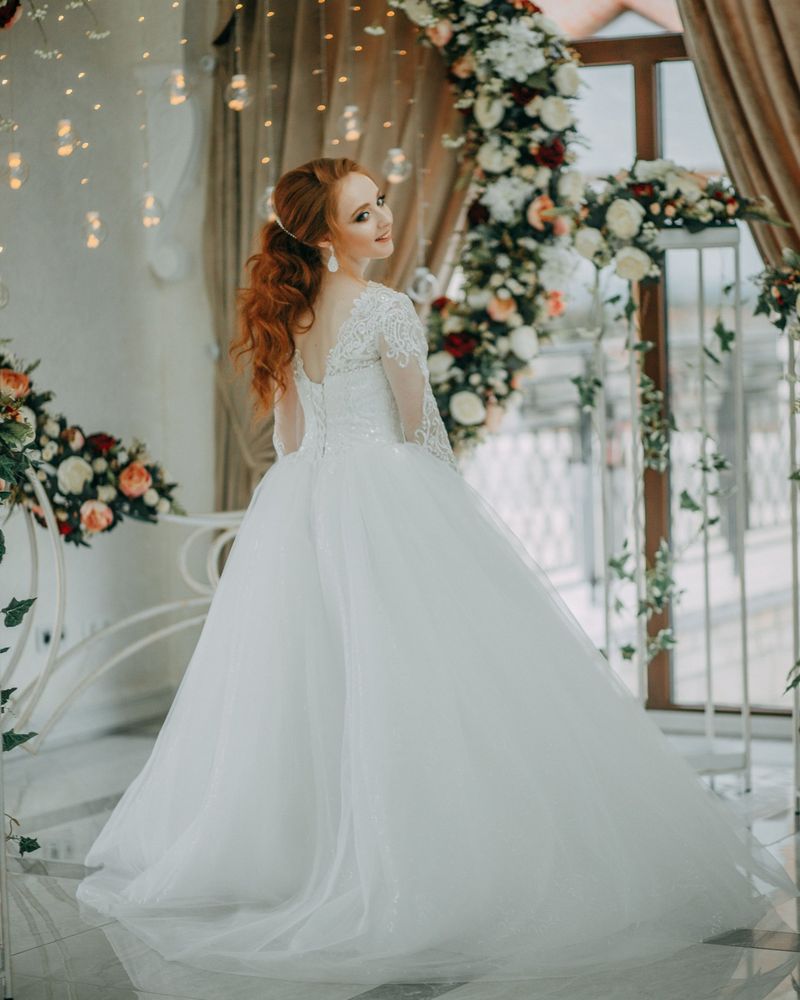 Red-haired bride glancing over her shoulder in a lace and tulle gown, with a flower arch and fairy lights behind her