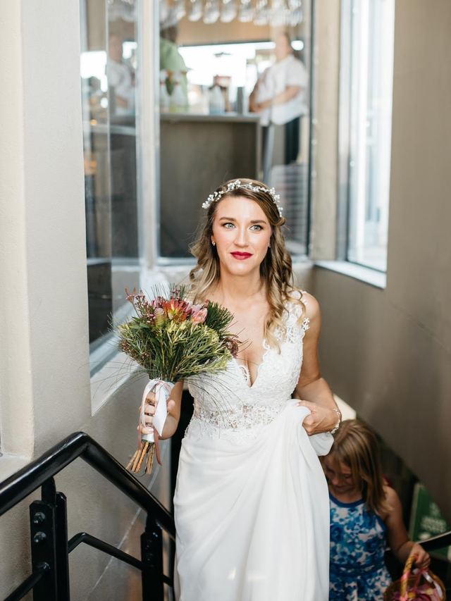 Bride in a delicate hair vine and red lipstick walking up a staircase with a protea bouquet