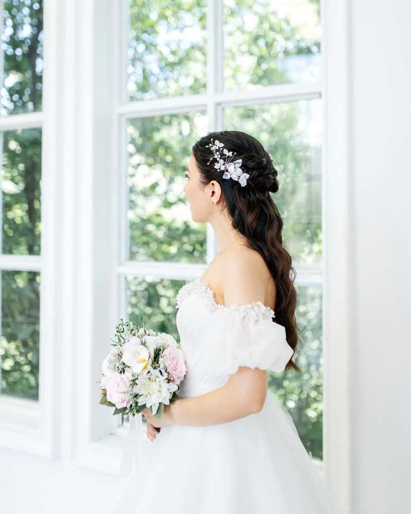 Bride in profile with a half-up twisted hairstyle, a leaf hairpiece and a pink and white bouquet by a bright window