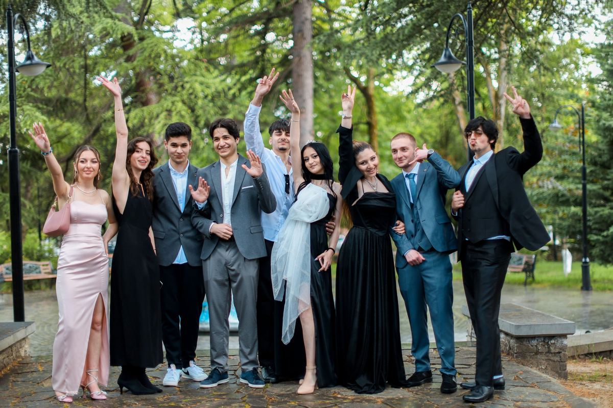 A group of friends in formal dresses and suits standing together outdoors, laughing and waving