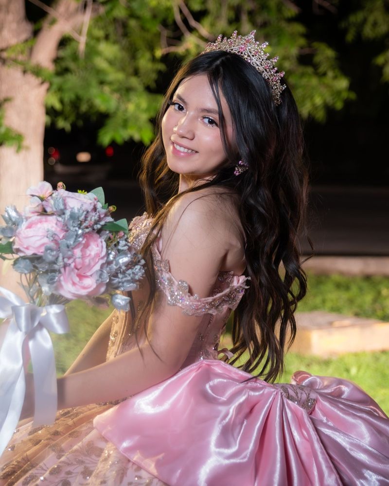 Girl with long wavy hair and a tiara, smiling over her shoulder in a pink sparkly gown, holding a pink bouquet
